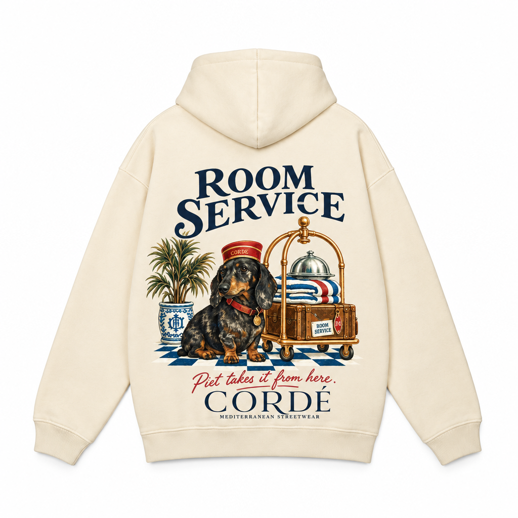 CORDÉ Cream hoodie, back: Piet in a bellboy hat beside a luggage trolley.